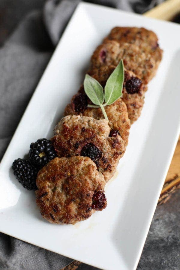 Six blackberry homemade sausages lined up on a white serving tray topped with basil and fresh blackberries