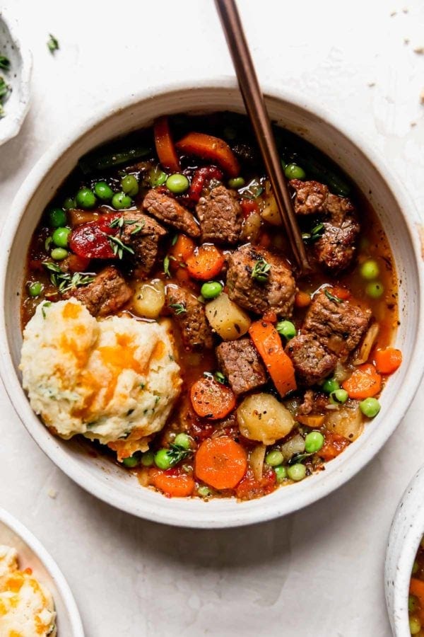 A hearty bowl of thick and chunky vegetable beef stew in a white bowl with a cheddar biscuit on the side.