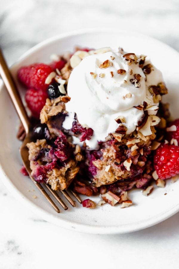 A large serving of mixed berry baked oatmeal on a speckled plate topped with whipped cream, fresh berries, and crushed nuts.