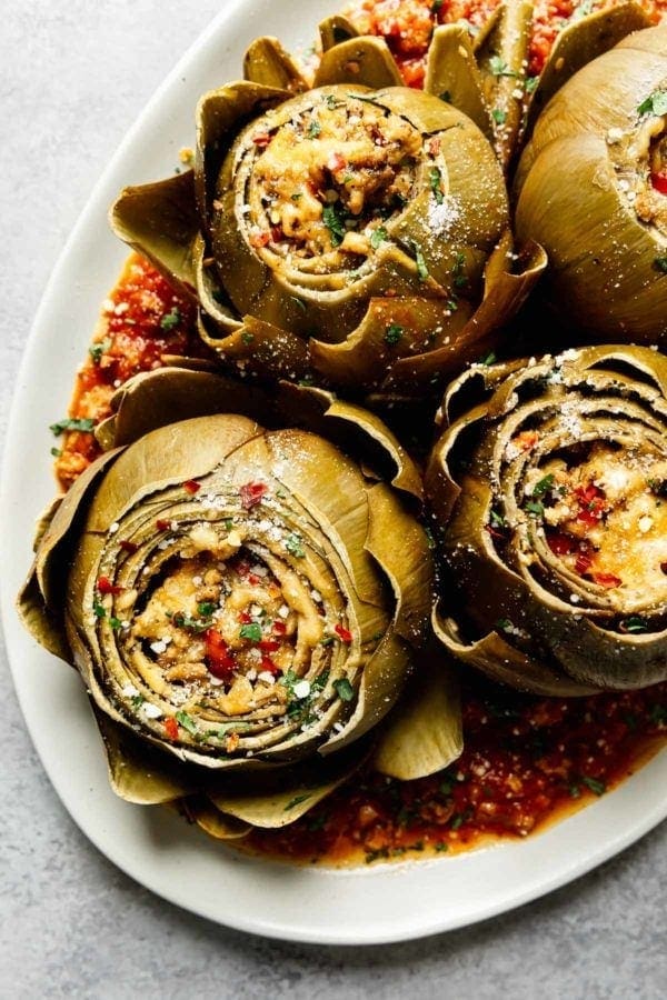 A platter full of Italian stuffed artichokes sitting in marinara sauce and topped with parmesan cheese