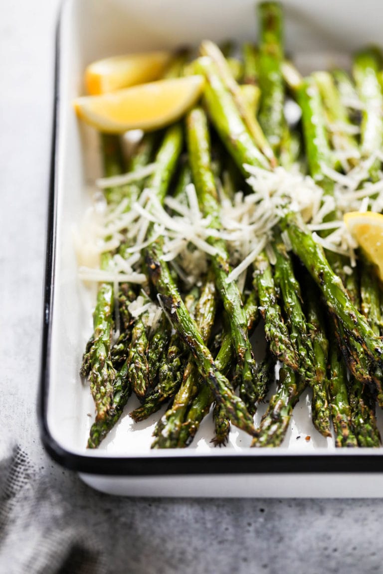Close up view of a serving pan filled with grilled asparagus topped with parmesan cheese and garnished with lemon wedges. 