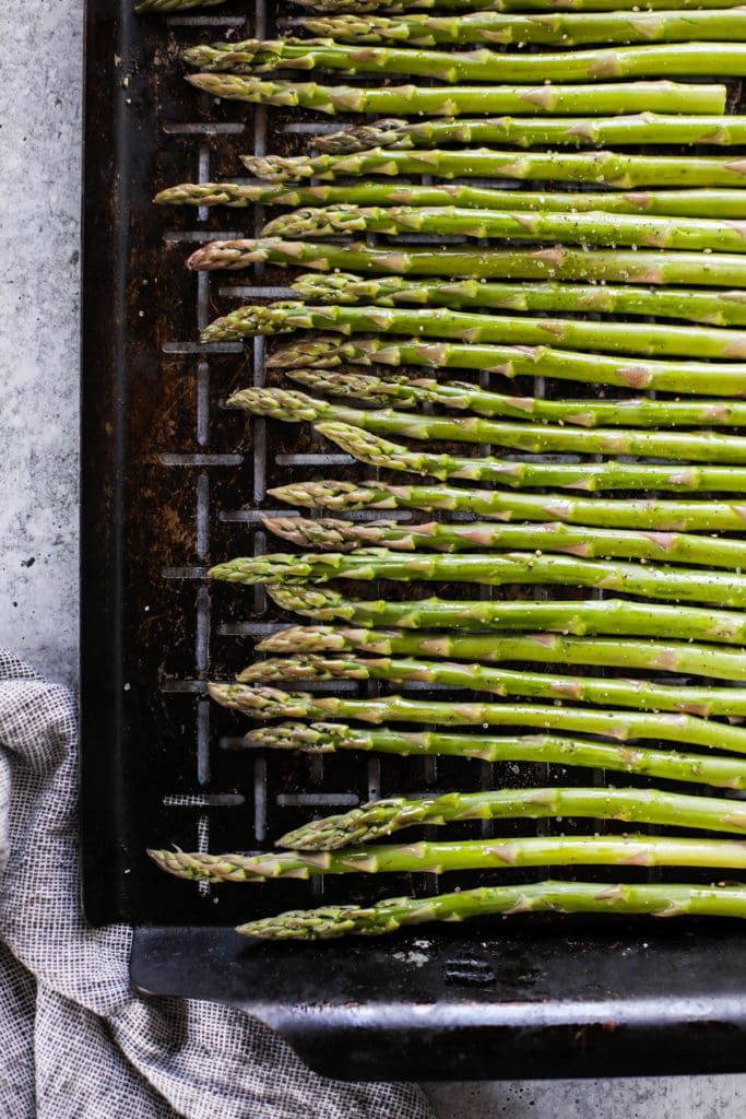 Overhead view of asparagus stalks ready for roasting on a pan topped with sea salt and pepper. 