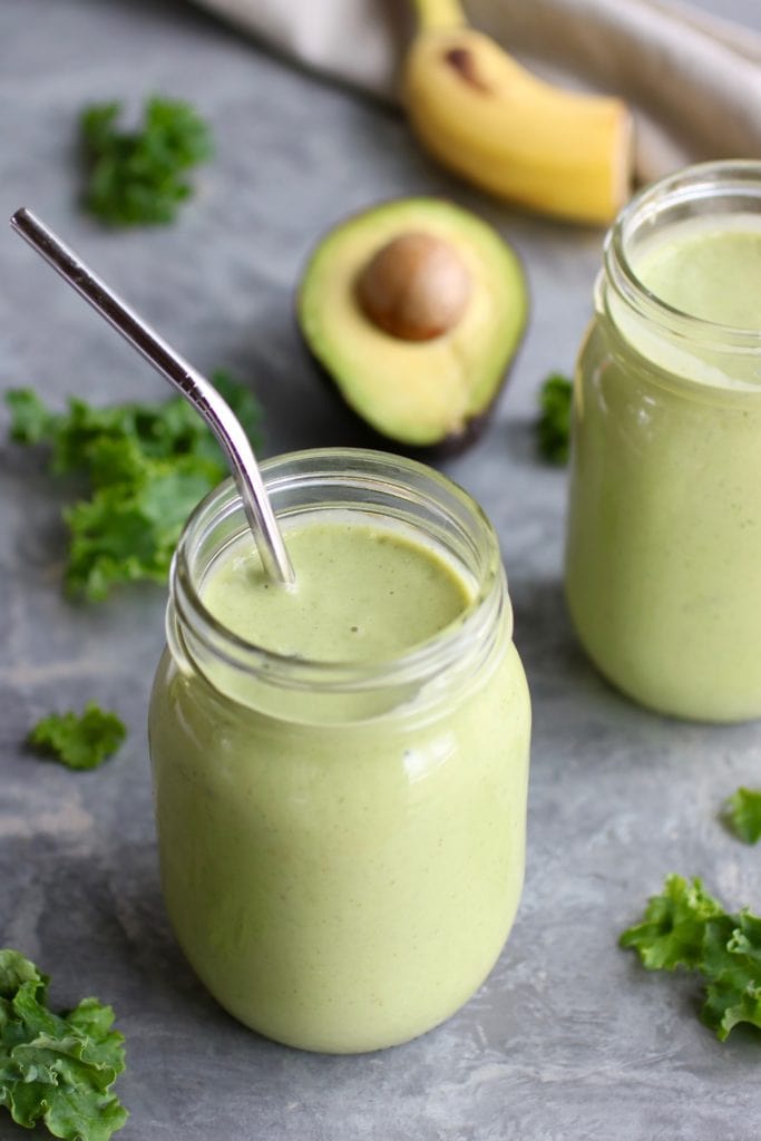 A mason jar filled with healthy green smoothie with fresh kale and avocado scattered around