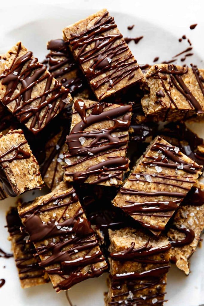 A pile of mini peanut butter protein bars cut into small squares topped with chocolate drizzle and sea salt.