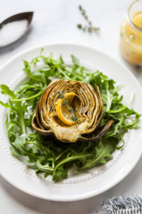 A roasted artichoke atop a bed of arugula topped with a thin lemon slice and fresh herbs
