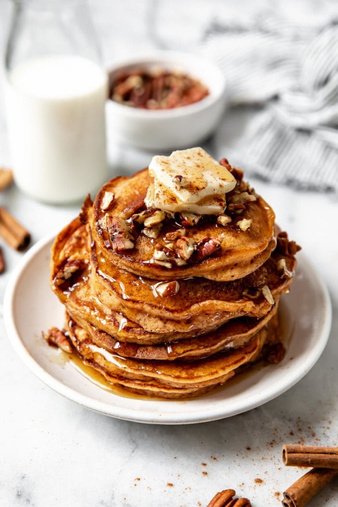 A tall stack of fluffy sweet potato pancakes on a speckled plate topped with a pad of butter, pecans, and maple syrup.