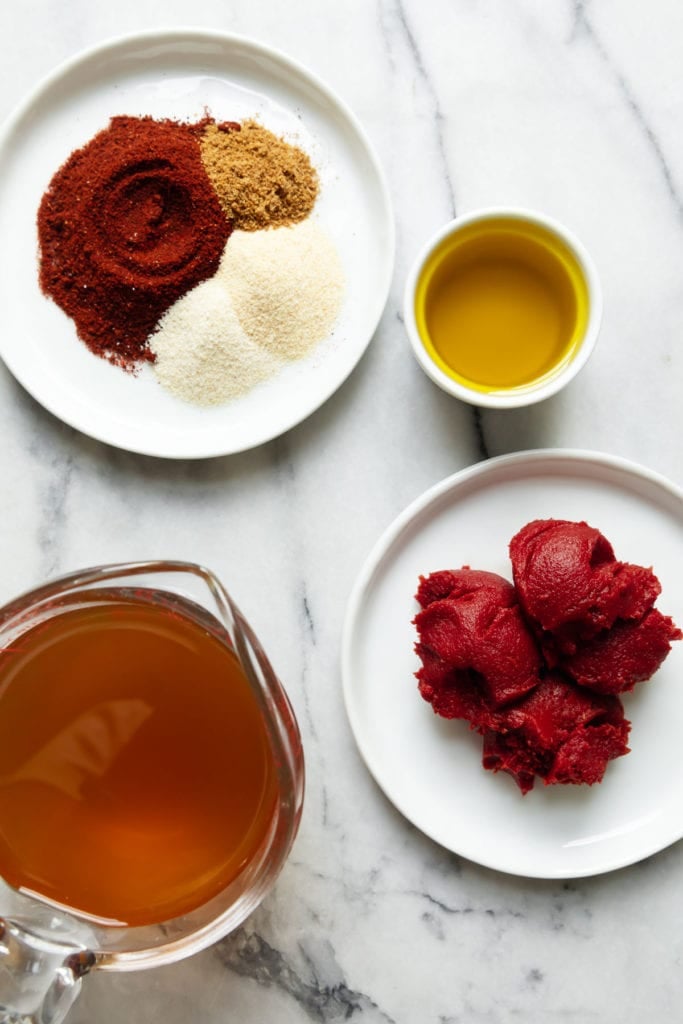 All ingredients for homemade enchilada sauce on small plates and measuring cups on a marble countertop