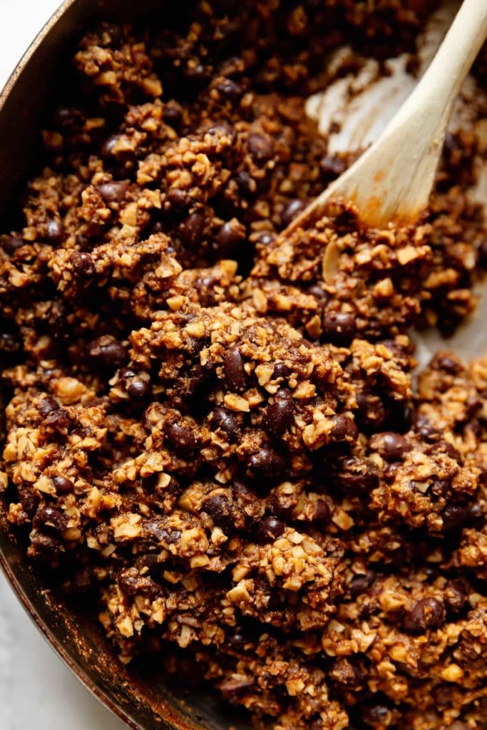 The vegan filling of cashews, walnuts, and black beans in a skillet being stirred with a wooden spoon.