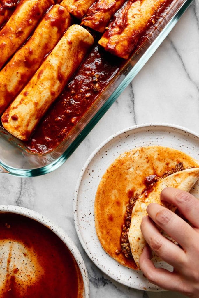 A vegan enchilada being rolled up on a speckled plate with a baking dish filled with enchiladas near