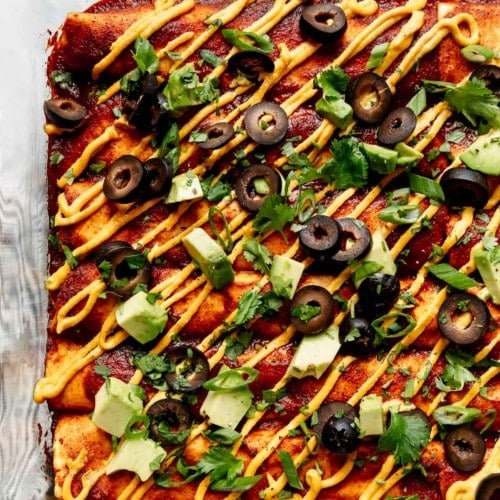 Vegan enchiladas lined up in a baking dish topped with homemade queso, avocado chunks, and black olives.