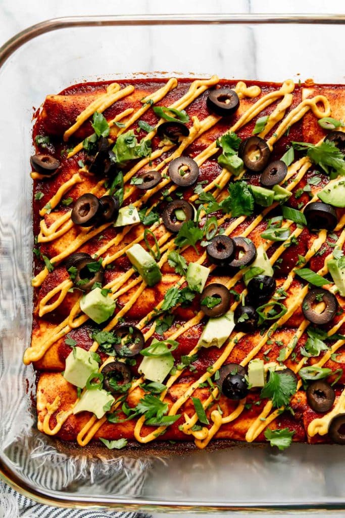 Vegan enchiladas lined up in a baking dish topped with avocado chunks, black olives, and homemade enchilada sauce