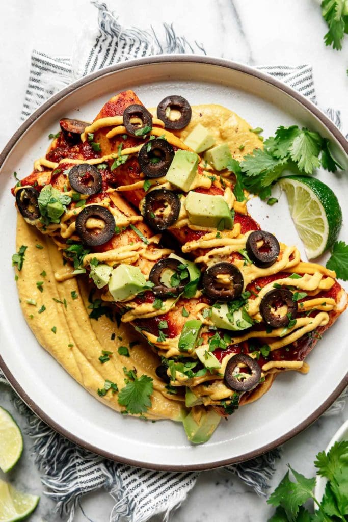 Two vegan enchiladas plated on top of a swirl of homemade queso and topped with cilantro, black olives, and avocado chunks