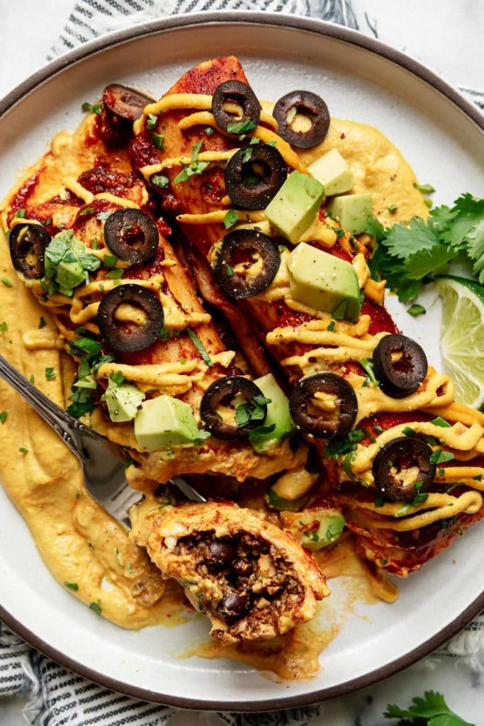 Two vegan enchiladas plated on top of a swirl of queso with a piece of enchilada on a fork to show texture