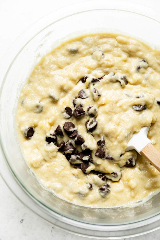 Creamy banana bread batter in a clear mixing bowl with a spatula folding in dark chocolate chips.