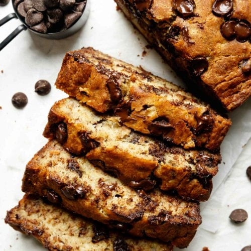 Several thick slices gluten free banana bread with melty chocolate chips cut from loaf, on white parchment.