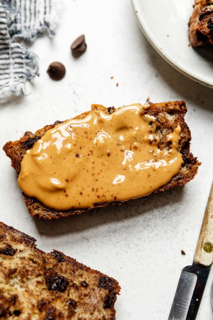 A thick slice of banana bread with chocolate chips topped with a slathering of creamy peanut butter. 