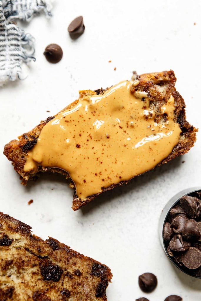 A slice of gluten-free banana bread with a slathering of creamy peanut butter with a large bite taken out of the corner