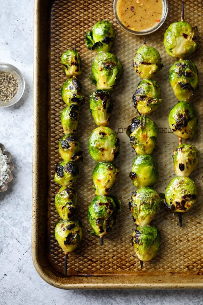 Grilled Brussels sprouts on skewers laying on a baking sheet with maple-mustard sauce in a bowl