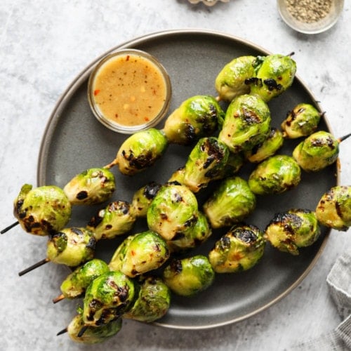 Grilled Brussels sprouts on skewers on a black plate with maple-mustard glaze drizzled on top