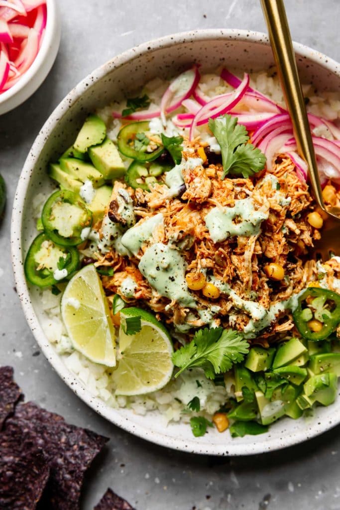 Instant Pot Salsa Chicken in a speckled white bowl served with cauliflower rice, avocado, cilantro, red onion, jalapeño and topped with cilantro-lime crema.