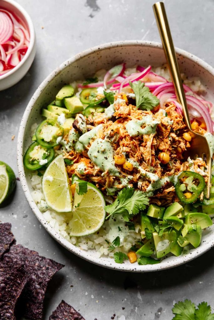 Instant Pot Salsa Chicken in a speckled white bowl served with cauliflower rice, avocado, cilantro, red onion, jalapeño and topped with cilantro-lime crema.
