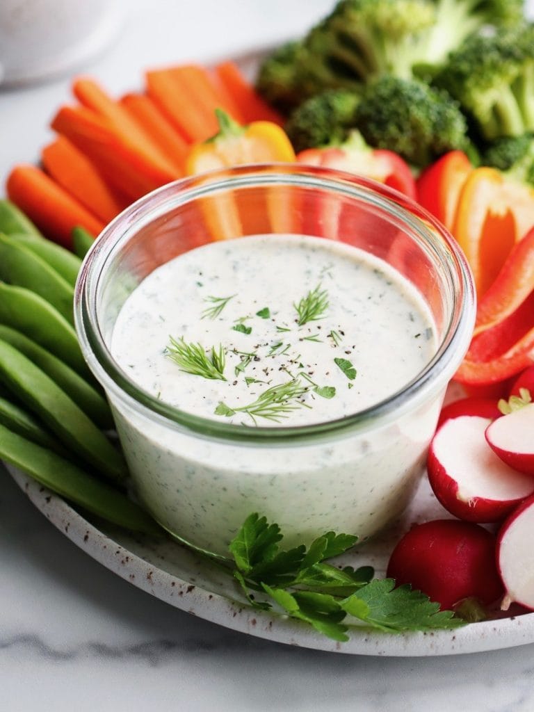 Dairy free ranch dressing in small glass jar on stone platter surrounded by fresh cut vegetables