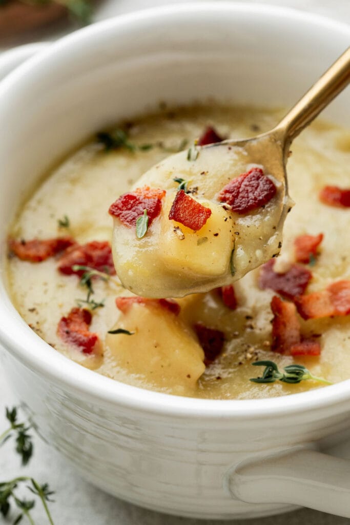 A spoonful of creamy potato leek soup garnished with crisp-cooked bacon, fresh thyme leaves, and cracked black pepper. 