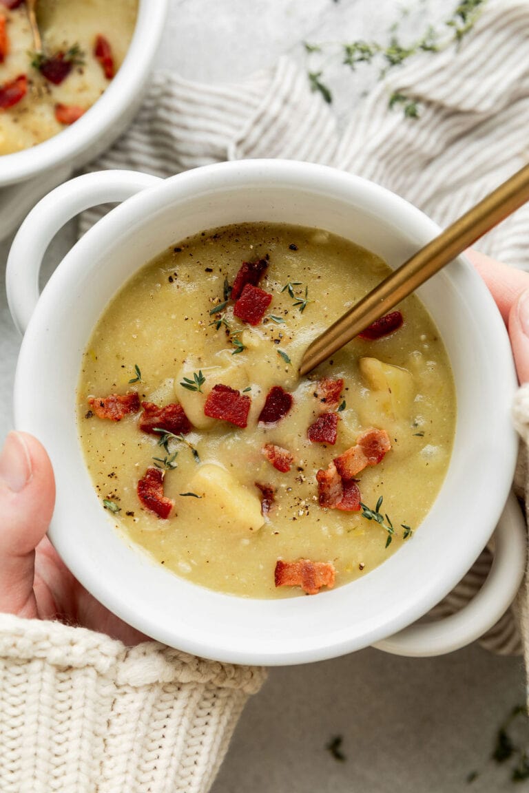Two hands cradling a small white bowl of creamy potato soup with leeks and bacon.