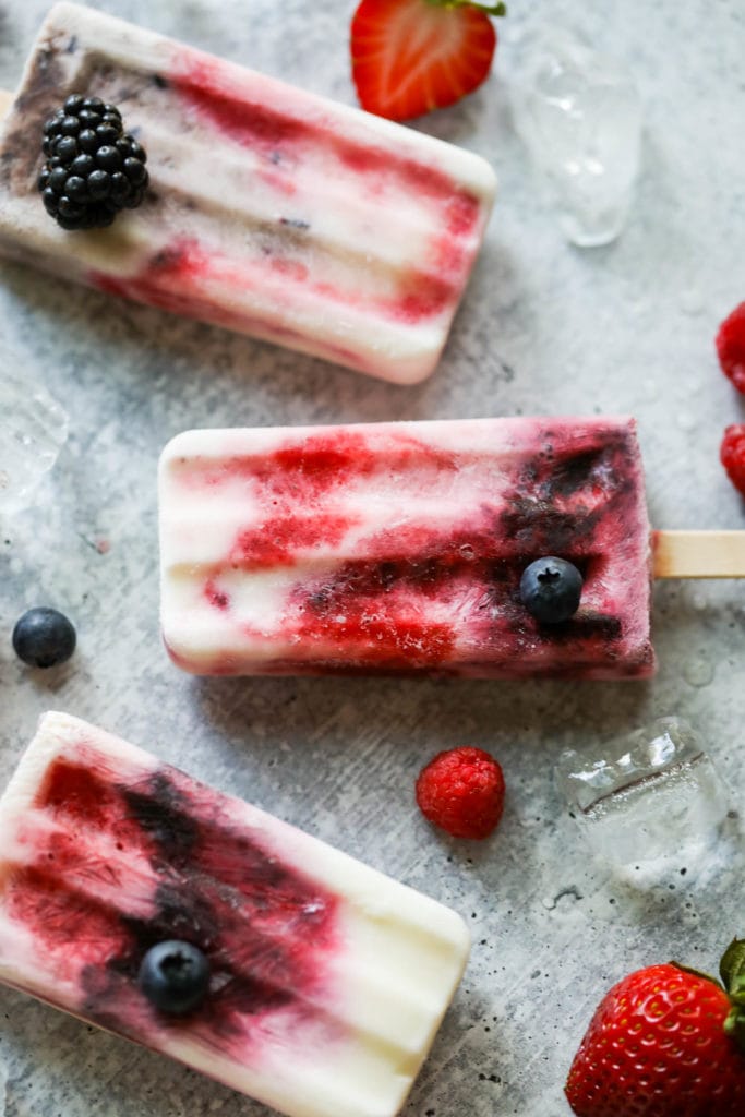 Three homemade creamy yogurt popsicles with berry swirls laying on a countertop with ice cubes and fresh berries scattered around