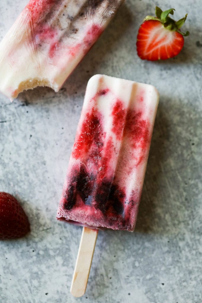 Creamy yogurt popsicles with berry swirls on a counter with fresh berries scattered around