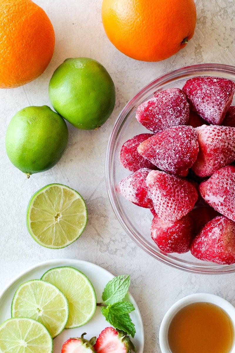 Frozen strawberries in a glass bowl with fresh limes and oranges nearby.