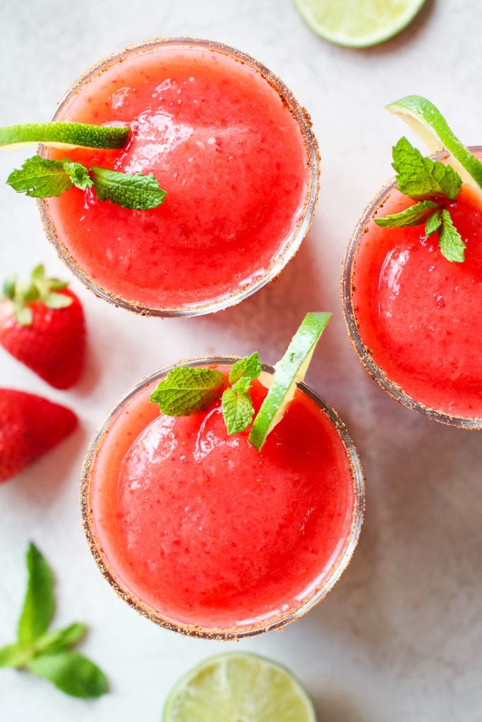 Three frozen strawberry margaritas in short glasses garnished with lime slices and fresh mint
