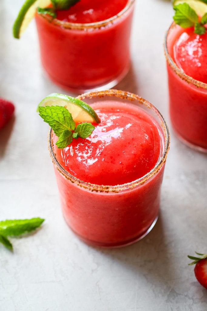 Three frozen strawberry margaritas in short glasses with coconut sugar on the rim and a lime and mint sprig on the edge