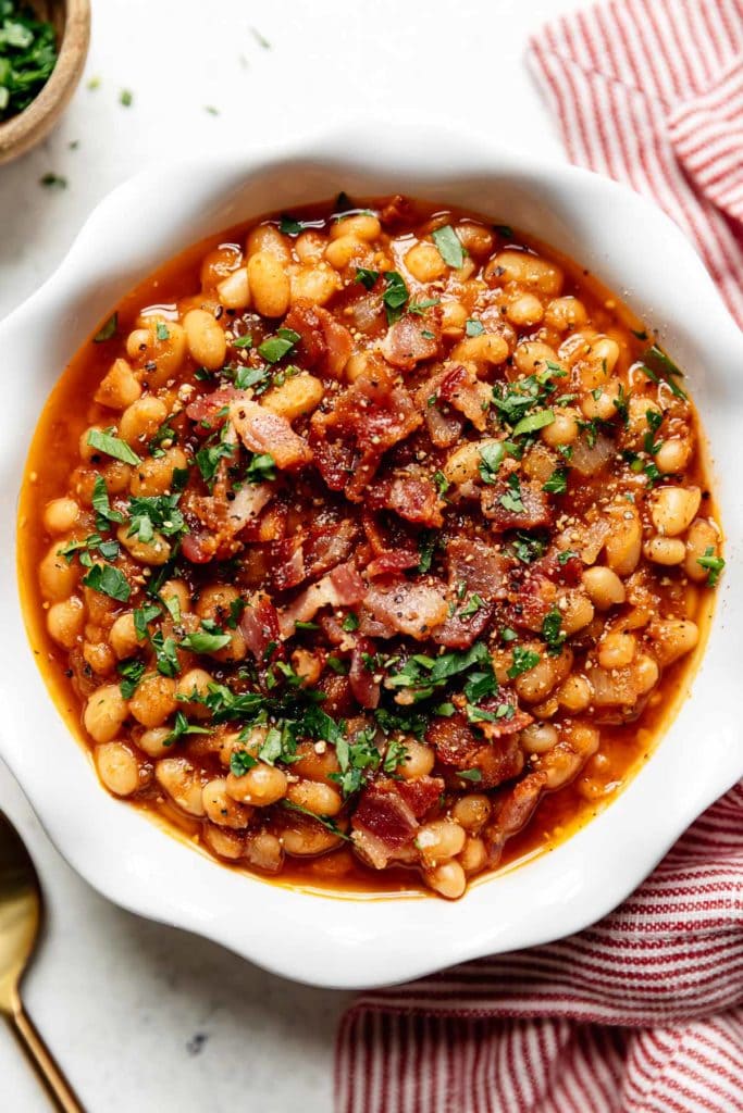 Overhead view of instant pot baked beans in a scalloped edge white bowl topped with bacon crumbles and fresh herbs. 