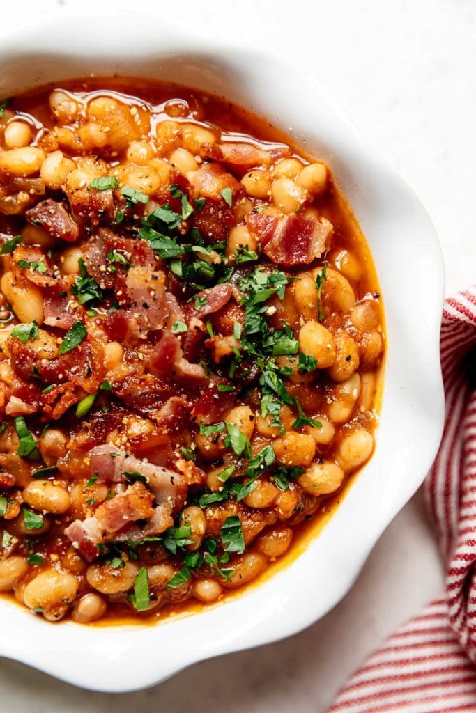 Close up overhead view of instant pot baked beans in a scalloped edge white bowl topped with crumbled bacon and fresh herbs. 