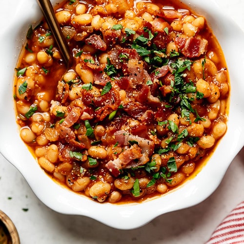 Overhead view of instant pot baked beans served in a scalloped edge white bowl topped with crumbled bacon and fresh herbs.