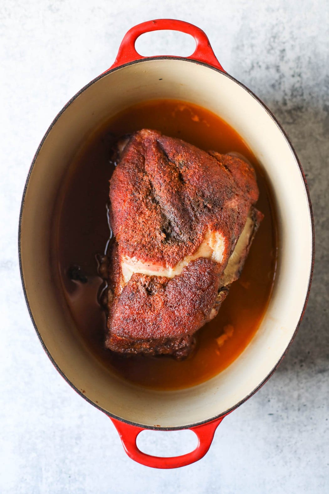 Smoked pork butt in a red dutch oven soaking in liquid ready to return to the grill