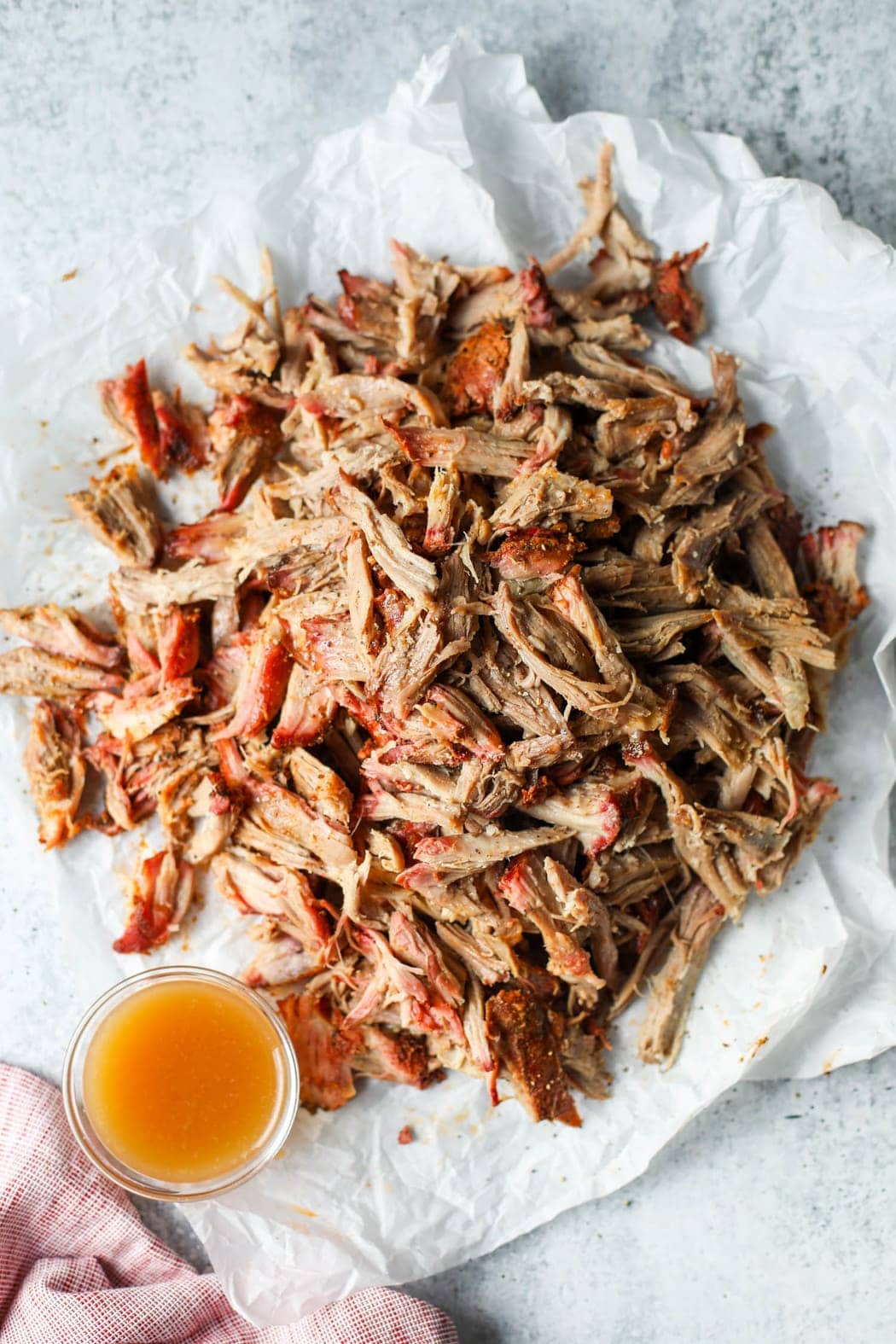 Smoked pork butt shredded, laying in a heap on a white towel with a small jar of apple cider vinegar on the side