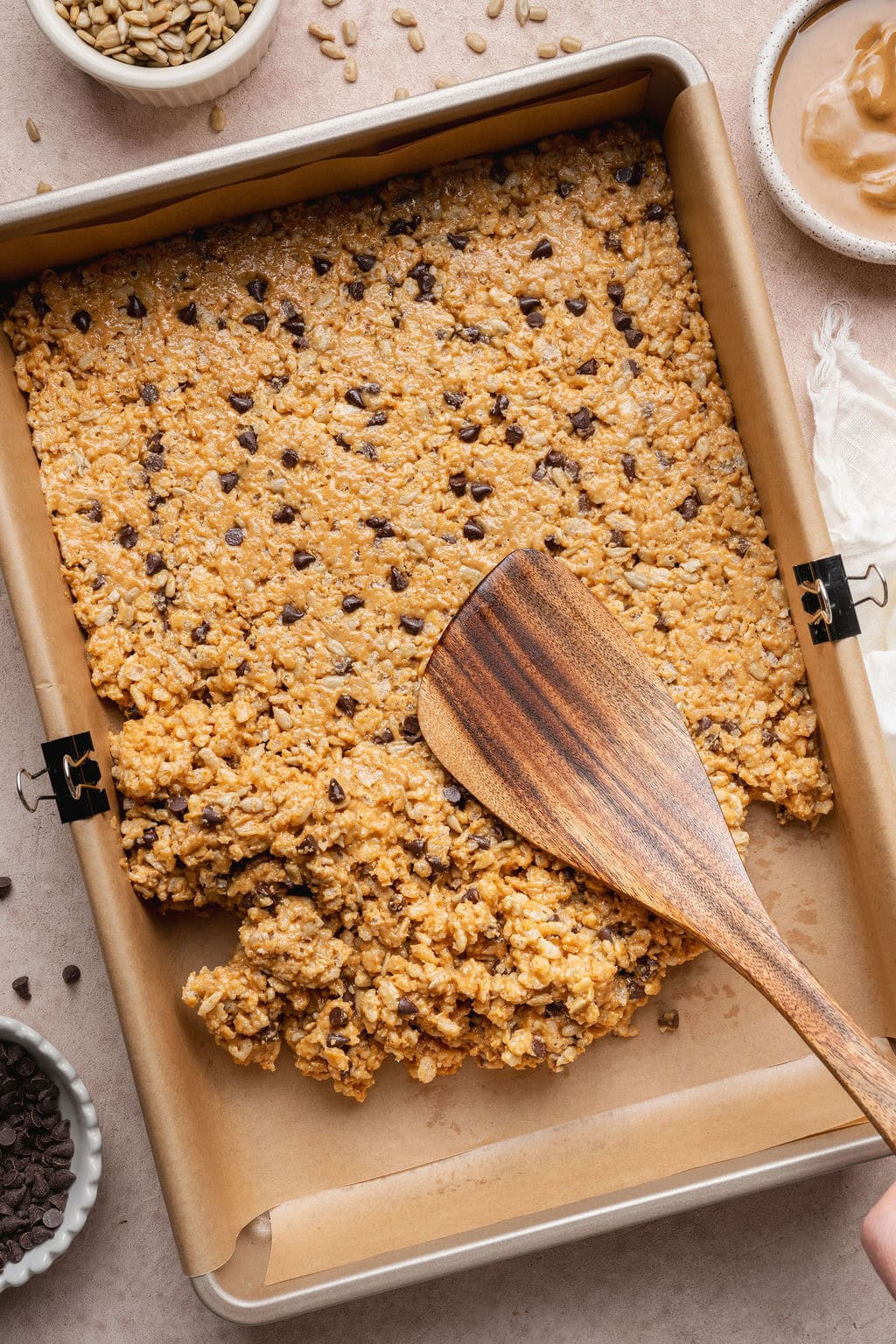 A wooden spatula spreading homemade Clif bar batter evenly over a parchment paper-lined baking sheet.