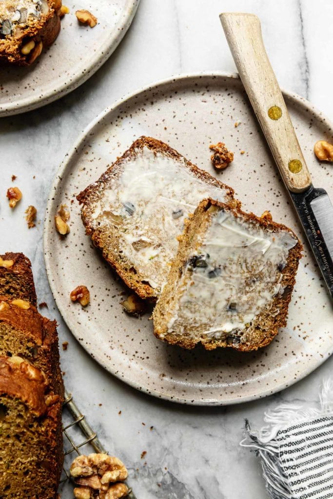 A thick slice of gluten free zucchini bread cut in half with a thin layer of butter on top plated on a grey speckled plate