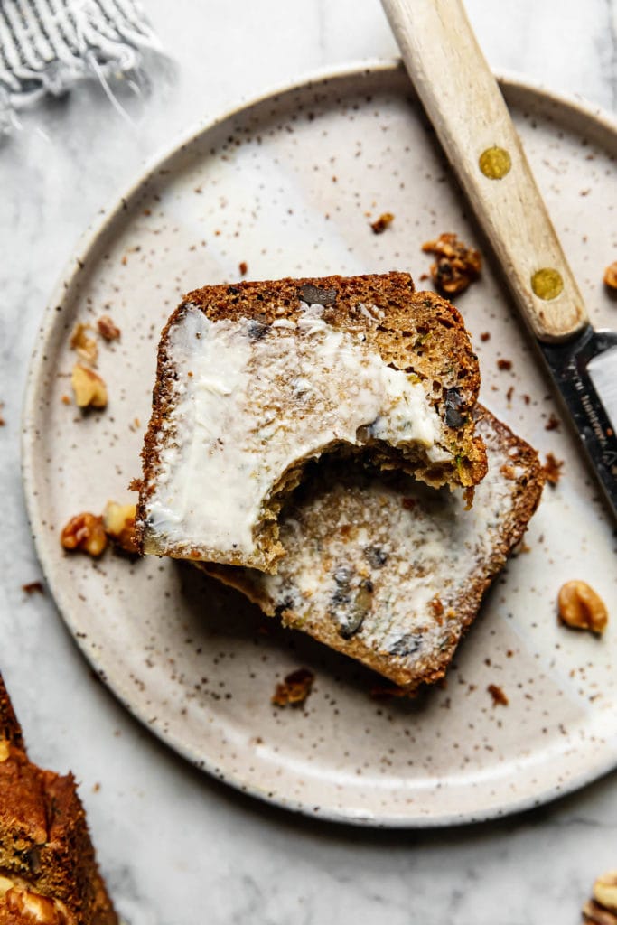 A thick slice of zucchini bread cut in half with a bite taken out of it topped with a thin layer of butter on a speckled grey plate