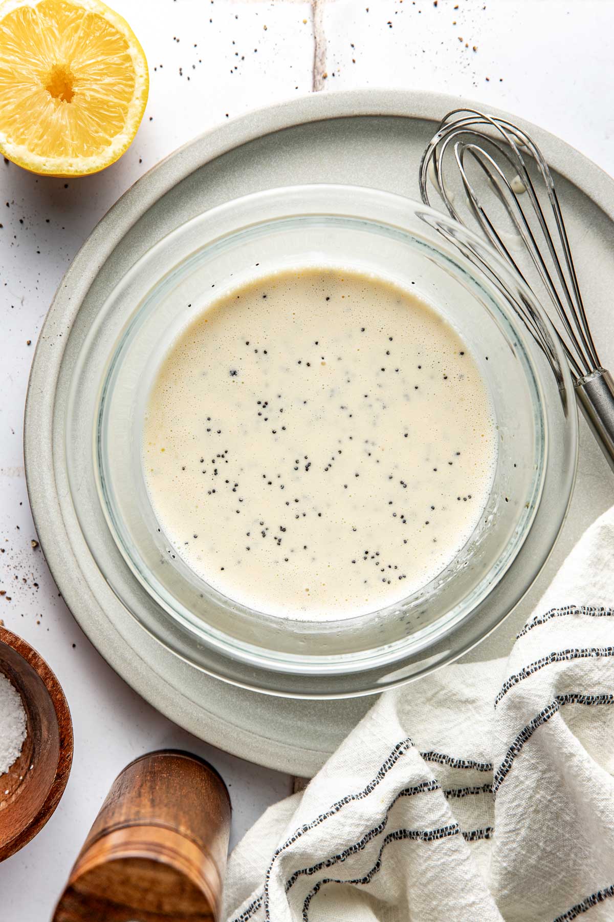 Overhead view of a glass bowl of poppy seed dressing next to a metal whisk and lemon wedge.