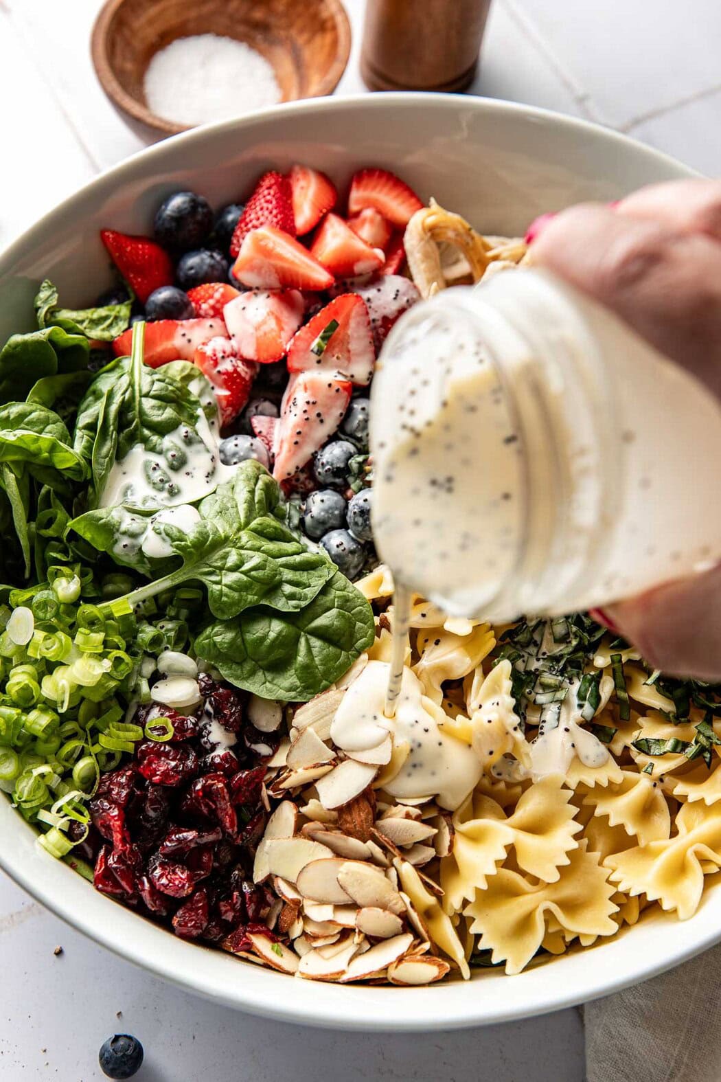 Close up view of a hand pouring a jar of poppy seed dressing on a bowl of fresh berries, spinach, and bow tie pasta.
