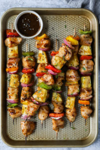 Grilled pineapple chicken kebabs lined up on a baking sheet with a small bowl filled with teriyaki sauce