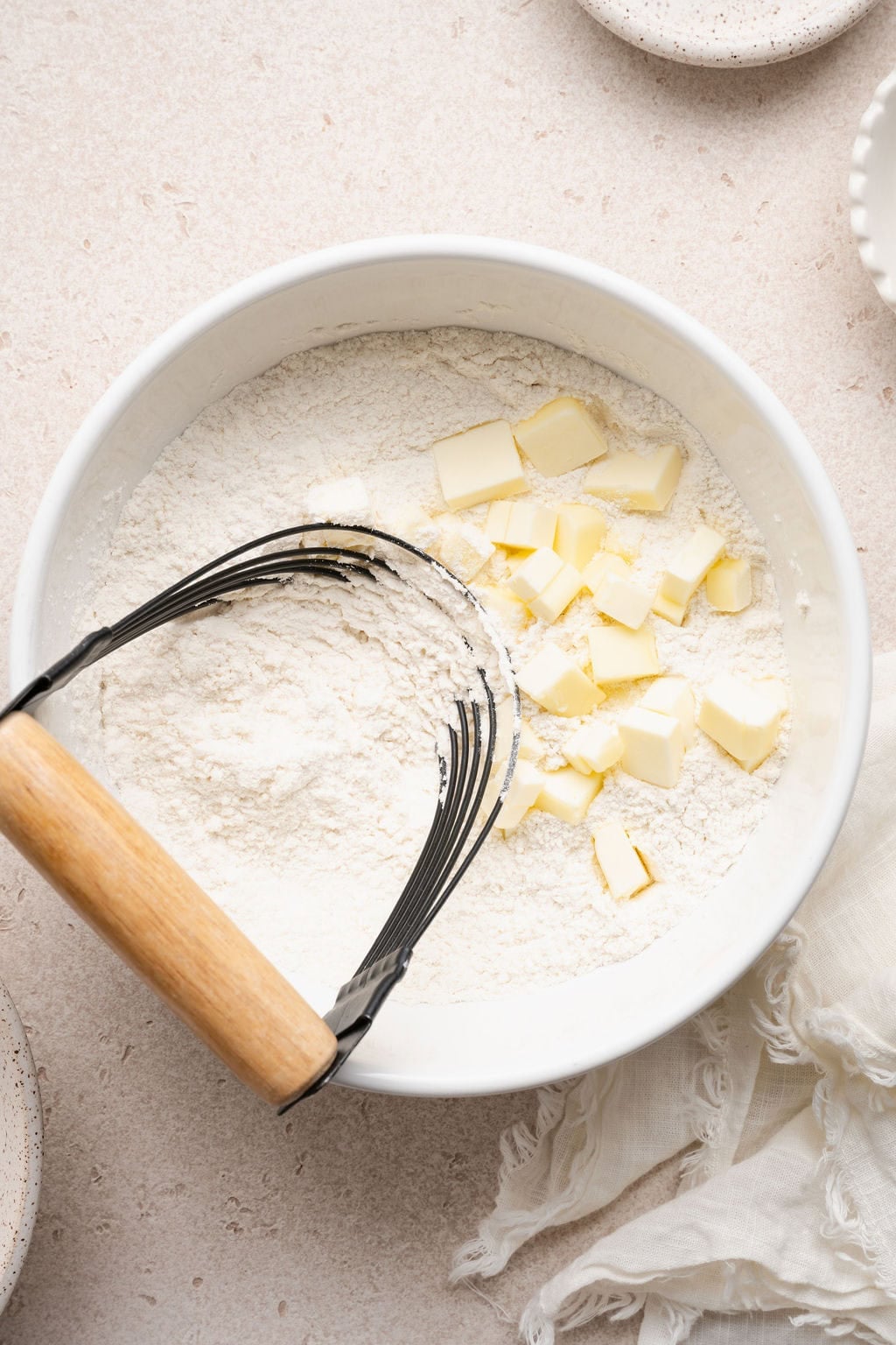 Overhead view of a white bowl of flour and cold cubed butter ready for combining with a pastry cutter on the side. 