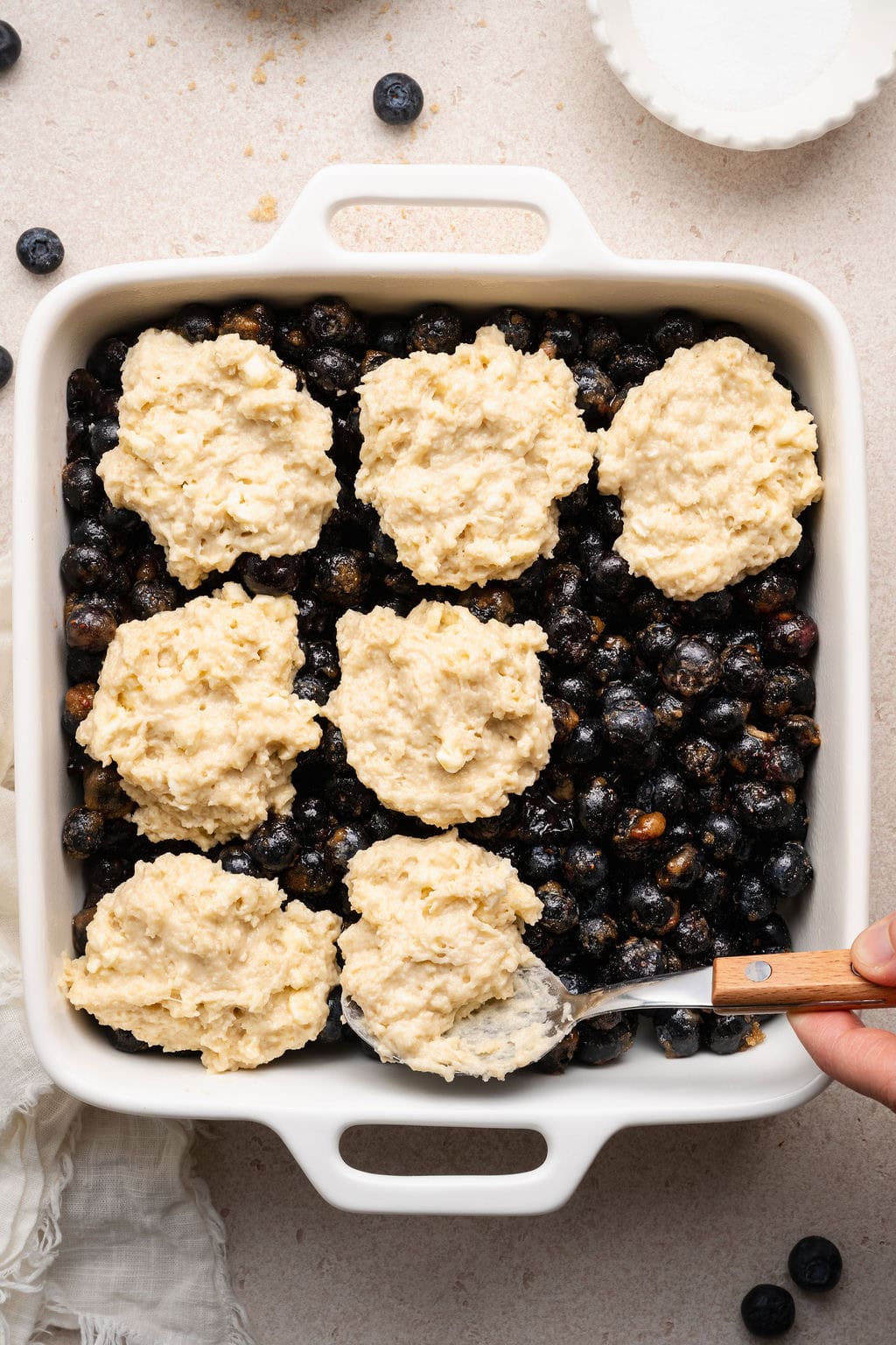 Overhead view of a baking dish filled with Blueberry Cobbler filling and a hand putting dough dollops on top. 