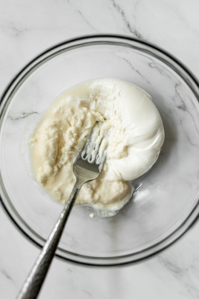 Fresh burrata being mashed with a fork in a clear bowl to show the creamy middle