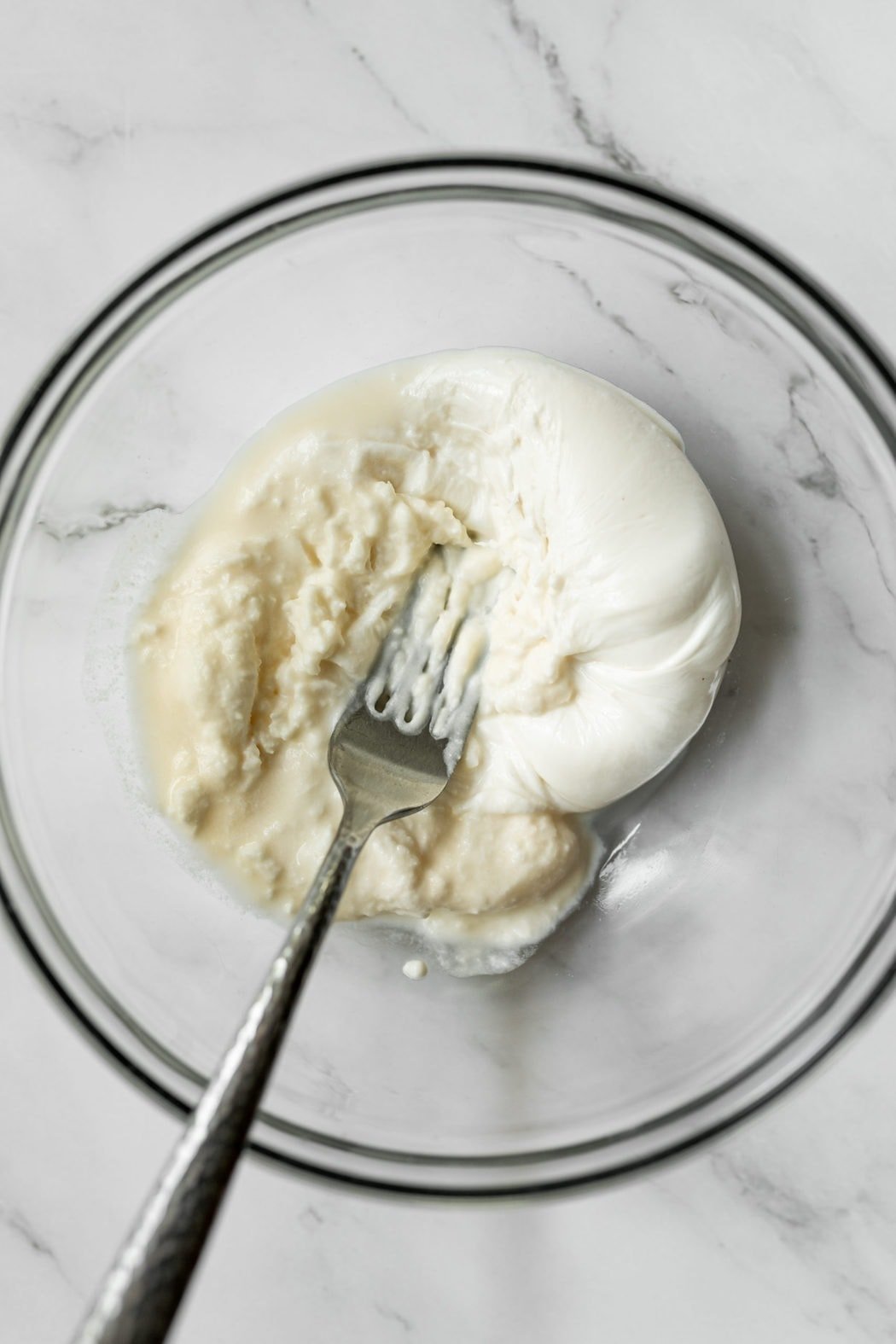 A ball of burrata being mashed with a fork in a mixing bowl