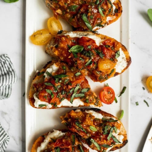 Slices of bruschetta topped with burrata, tomatoes, and basil on a white platter