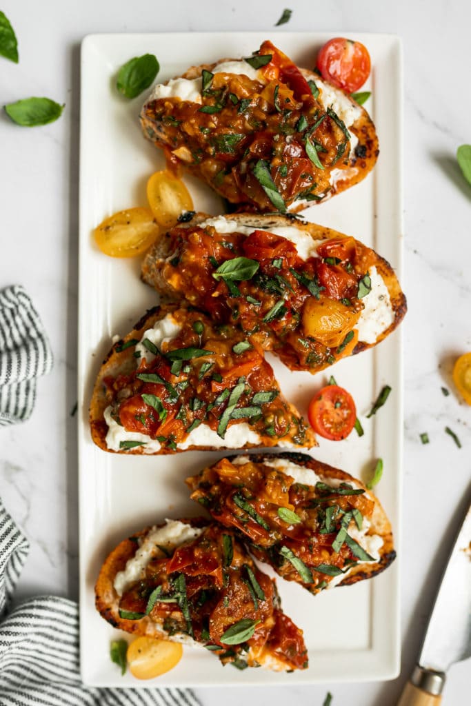 Bruschetta topped with burrata, tomatoes, and fresh herbs on a white serving tray
